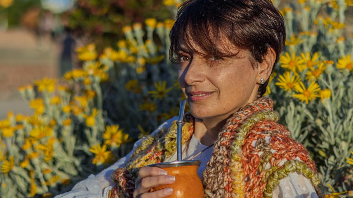 Woman with handcrafted scarf sipping yerba mate outdoors in Montevideo. Vibrant yellow flowers in full bloom.