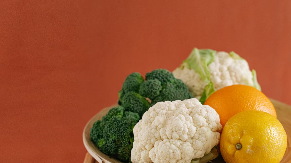 Close-up of fresh broccoli, cauliflower, lemon, and orange on a wooden plate with an earthy background.